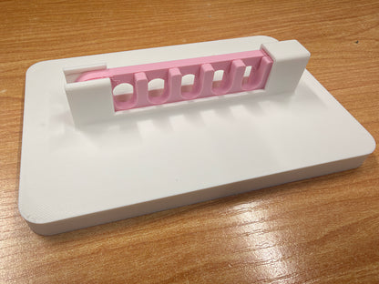 A pink plastic yarn guide with five curved slots, resting on a white plastic storage tray over a wooden tabletop.