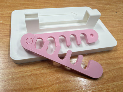A pink plastic yarn guide with five curved slots, resting on a white plastic storage tray over a wooden tabletop.